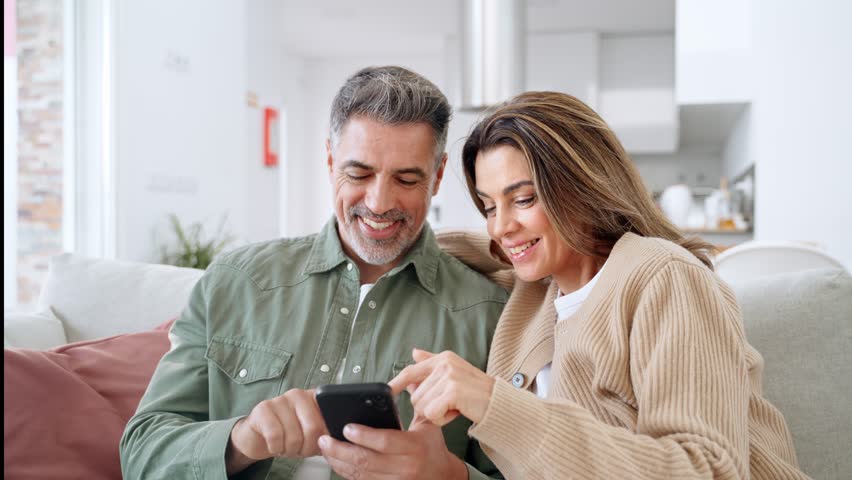 Happy older mature family couple, middle aged man and woman looking at cell phone using smartphone mobile technology device together sitting at home relaxing on couch doing online shopping. - Powered by Shutterstock - Get 15% off with code: PIKWIZARD15