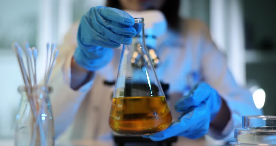 Scientist in blue gloves holds a tilted round-bottomed flask with a yellow liquid Research oil and petroleum products