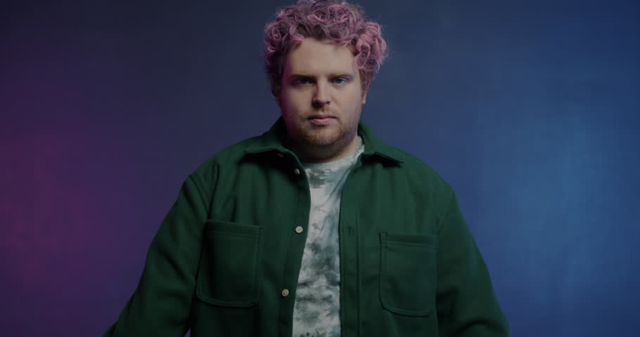 Slow motion portrait of creative young man moving hands with neon light sticks and looking at camera on purple background. People and illumination concept.