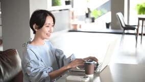 Smiling, beautiful Asian (Japanese) woman working at a computer in an office or cafe. Video of her talking to a client online.