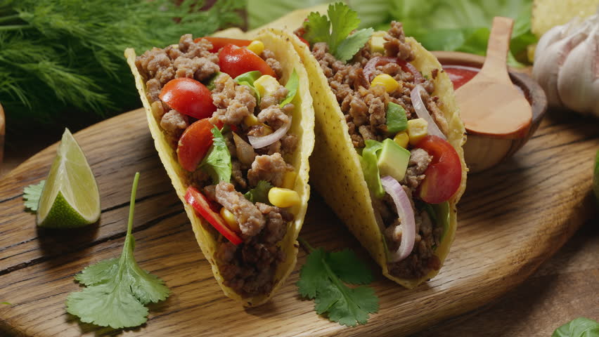 Two meat tacos on wooden board surrounded with fresh herbs and vegetables – traditional Mexican food spinning slowly on clockwise.