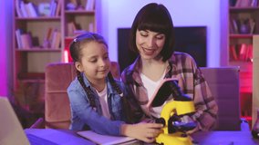 Happy little daughter and her mother sitting together at table and looking into electron microscope at evening time. Two caucasian women wearing casual clothes making scientific research at home. - Powered by Shutterstock - Get 15% off with code: PIKWIZARD15