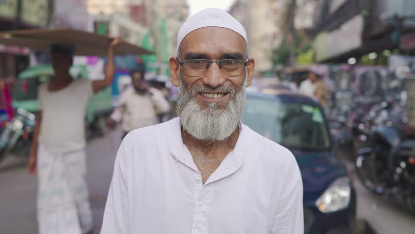 An old or aged happy Indian Asian Muslim bearded man or male in traditional attire and eyeglasses standing outdoors looking at the camera and smiling on a busy market street or narrow road 