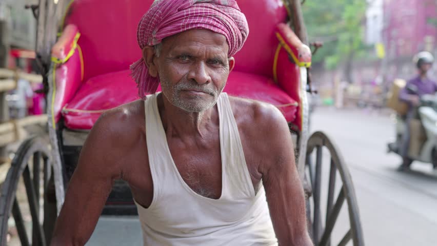 An elderly or old aged Indian Asian man or male traditional cycle rickshaw puller or driver wearing a head scarf looking or facing the camera on a busy market road or street