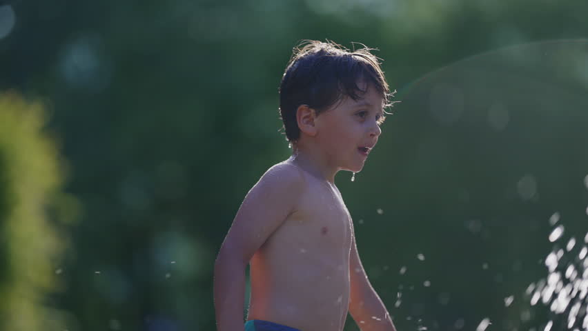 Little boy refreshing himself at public swimming pool water fountain in slow-motion 120fps. Child enjoying splashing water droplets during hot summer day