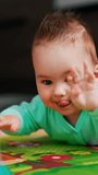 Funny cute baby with cute hairdo lies on the floor. Lovely child claps his hands on the floor and smiles. Blurred backdrop. Vertical video