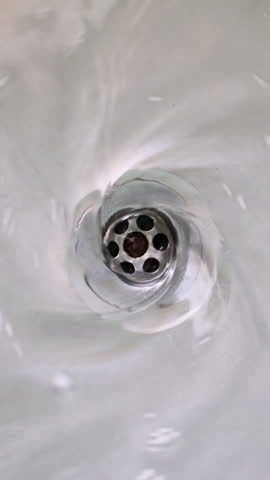 Water swirling down the drain hole of a stainless steel sink, close up. Vertical video.