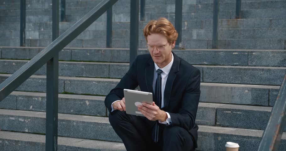 Suited man on steps using tablet tool for leisurely internet use Work pause with tablet offers digital escape. Engrossed in tablet he balances work with downtime blending connectivity and relaxation.