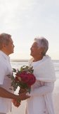 Vertical video of happy senior biracial couple kissing on sunny beach. healthy, active retirement beach holiday.