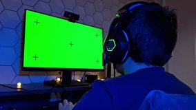 Rear view of a teenager boy with headphones game at a computer in a dark room with blue lighting