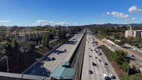 aerial footage of cars and trucks driving on the 210 Freeway at Lake Avenue surrounded by lush green trees, blue sky, clouds and buildings in the skyline in Pasadena California USA