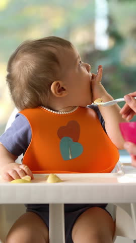 Mother feeding her baby son with fruit vegetable puree in baby chair. Vertical video. Royalty-Free Stock Footage #3474934357