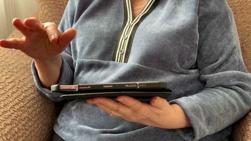 A stylish old woman sitting at a beige armchair looks for important information using an smart tablet at home. Close up