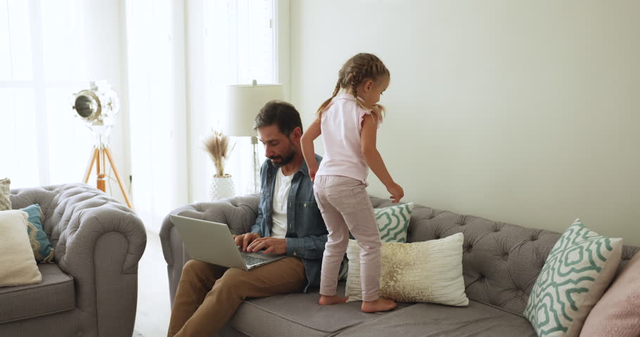Focused middle-aged man sitting on couch working on wireless computer, do business task, engrossed on remote job, his little 4s daughter disturbing daddy, jumping nearby. Telework, freelance from home Royalty-Free Stock Footage #3475158863