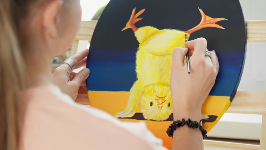 Young woman painter drawing yellow chicken on canvas using acrylic paints. Female artist works in studio. Drawing concept. Hobby. Canvas stands on easel. Close-up in 4K, UHD
