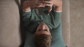 Portrait of thoughtful teenager Boy lying on sofa using smartphone. Scrolling social media, surfing internet at home alone. Serious child laying on coach in living room with phone. View from above