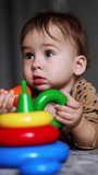 Adorable one-year old kid lies on the floor in the room. Baby holding parts of a toy pyramid in his hands. Blurred backdrop. Vertical video