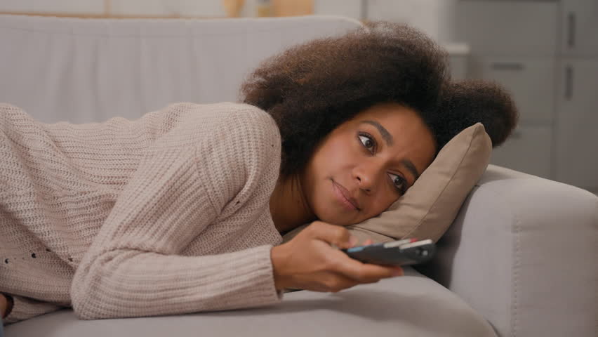 Bored dissatisfied upset African American woman lonely young girl lazy homeowner female lying on sofa couch searching TV channel program with remote control watching television boring movies at home Royalty-Free Stock Footage #3475543595