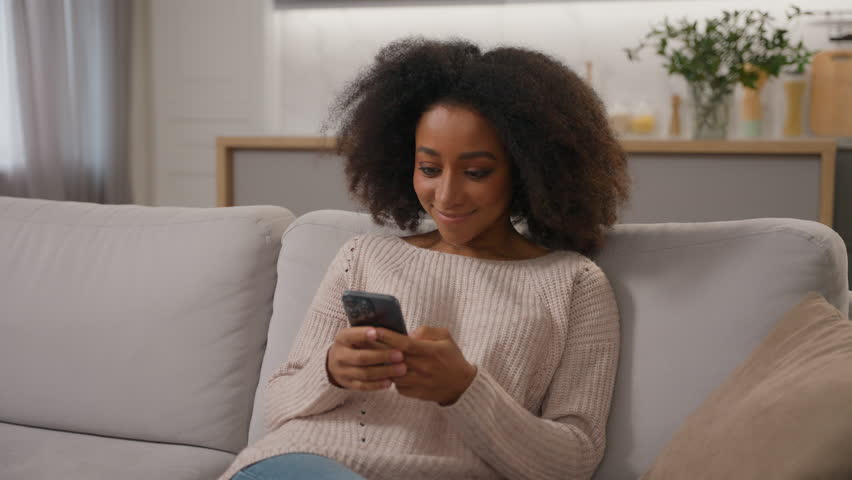 Happy African American girl smiling ethnic female woman using mobile phone cell gadget addicted chatting on smartphone shopping online on home sofa scrolling social media addiction influencer blogger Royalty-Free Stock Footage #3475554973