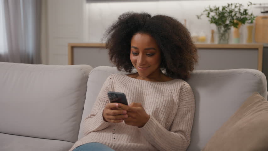 Happy African American girl smiling ethnic female woman using mobile phone cell gadget addicted chatting on smartphone shopping online on home sofa scrolling social media addiction influencer blogger
