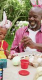Vertical video of happy african american grandfather and granddaughter painting eggs at easter. family spending time together at home.