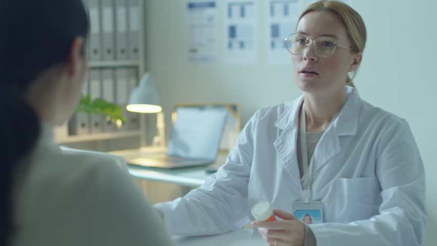 Beautiful female doctor in white coat explaining the dose and giving pills to female patient during medical consultation in clinic