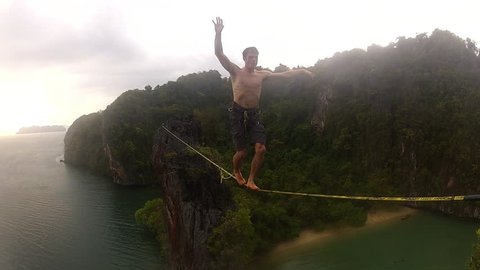 A brave and adventurous climber walks across a slackline over a bay in a tropical setting, with the ocean and jungle behind him, during the day