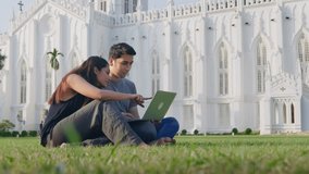 A young Indian happy good-looking male and female romantic couple sitting on the green lawn outdoors using a laptop to watch a funny video or movie smiling together. Relationship, affection concept