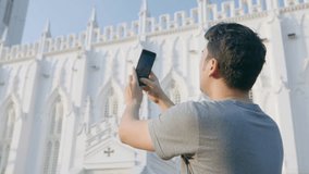A young Handsome Indian Asian man or guy is taking pictures or shooting a video vertically of an ancient Gothic architecture-style cathedral church on a mobile phone or smartphone camera in daylight.
