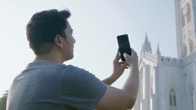 A young attractive Indian Asian man or male is taking photos or shooting a video of an ancient Gothic architecture style catholic church building on a mobile phone or smartphone camera in daylight.