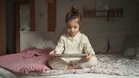 Cute girl sitting on on bed in bedroom watching something on tablet. Children's screen time.