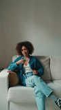 Vertical video, Young woman with curly hair wearing denim shirt looking at mobile phone while sitting on sofa in cozy living room