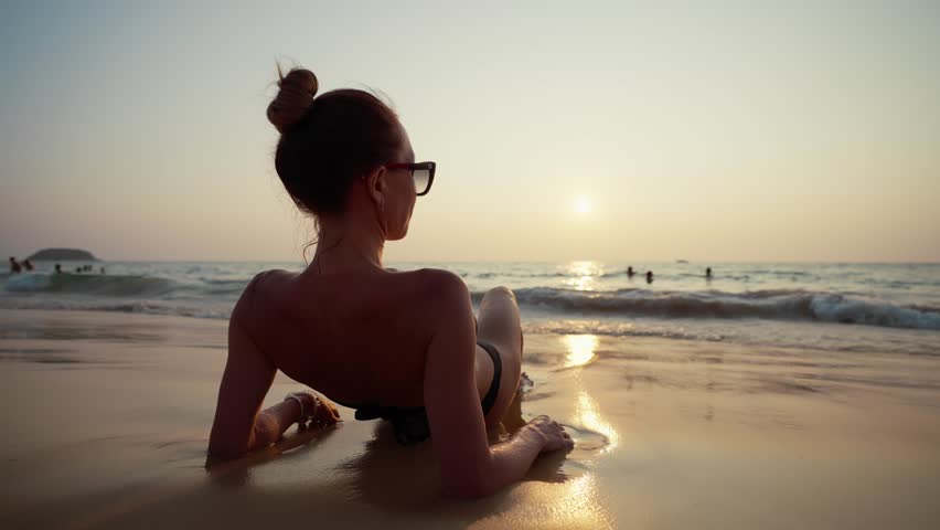 Blonde woman in black bikini at sunset. Girl lying on ocean beach in tropical paradise island with water waves, admire white sand in sunny weather back view. Beach resting, female travel, tourism.