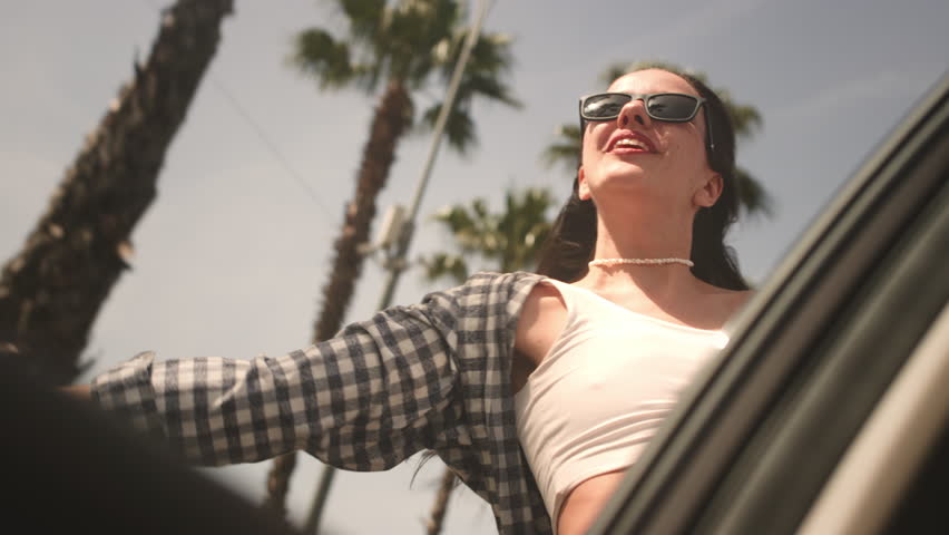 Excited woman enjoys the palm trees of seaside road. Girl leans out of the window of a rental car on a coastal highway on a sunny day. Hair in wind. A tropical vacation on the beach of exotic islands Royalty-Free Stock Footage #3476021339