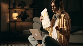 A Young Beautiful Woman Communicates Emotionally with her Colleagues in a Video Conference from Home. Girl quarrels and tears papers with printed graphs and tables. Backlit Warm Light