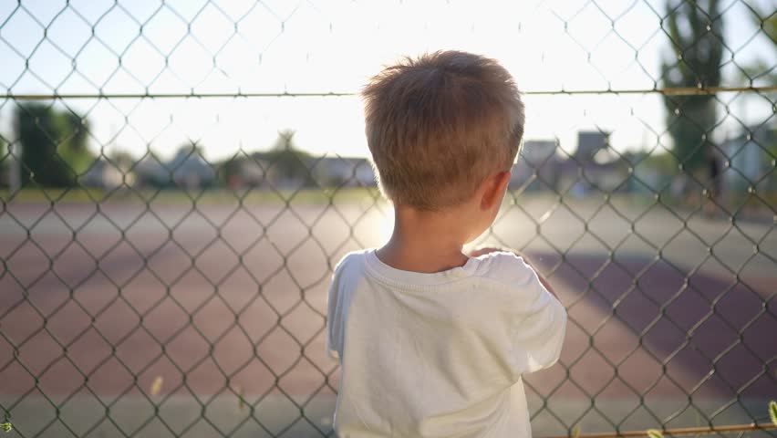 boy stands near the playground. concept of happy childhood and loving family. a child holds on to the net of a sports ground, court and glare of the sun lifestyle in the background