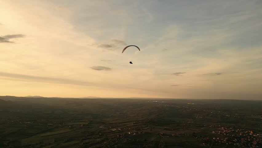Motorized paraglider flies low over 
 rural land in sunset.