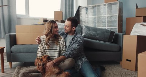 Boyfriend and girlfriend sitting on carpet near sofa with dog. Nice young Caucasian people smoothing Spaniel and admiring new apartment. Smiling.