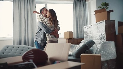 Nice Caucasian couple in love hugging and turning. Young people moving to new apartment. There are unpacked things, new sofa in living room.