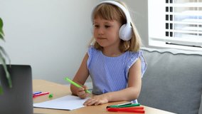 Junior schoolgirl with headphones waves hand to teacher at video lesson at home. Happy little student listens to online art class via laptop at desk