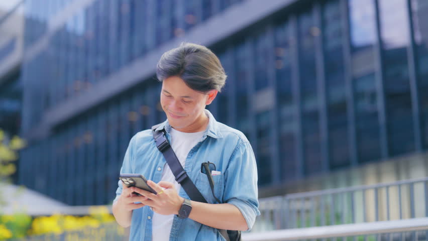 Happy smiling young Asian man using mobile smartphone outdoors on street in the city