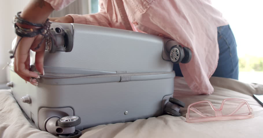 A young African American woman is packing a silver suitcase for vacation. She has a smartphone and pink sunglasses nearby, signaling travel preparations, slow motion, unaltered.