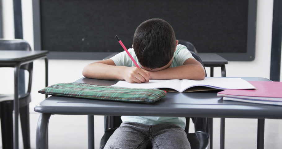 In a school classroom, a young biracial male student rests his head on his arms. He is wearing a light green t-shirt, focusing on a notebook with a pencil in hand, surrounded by textbooks.