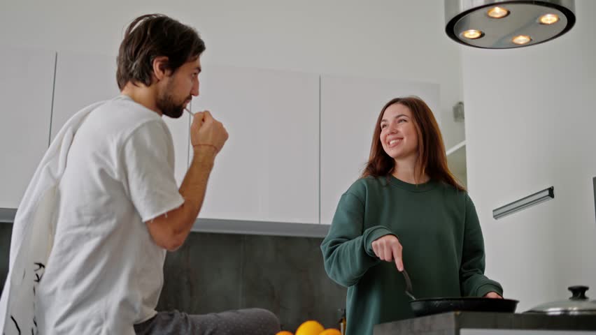 A brunette man with stubble and in a white T-shirt brushes his teeth while his brunette girlfriend in a green sweater makes breakfast in the kitchen in a modern apartment