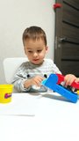 Toddler kid sits at desk focused on his toy. Adorable baby plays with plasticine and molding device. Vertical video.