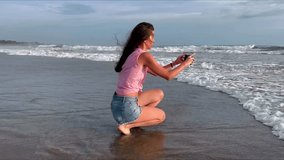 A beautiful girl with long black hair takes pictures of the ocean on her smartphone. A woman walks along the coast of a popular beach and shoots a video of foaming sea waves at sunset. High quality 4k