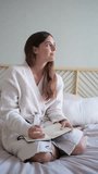 Pensive happy smiling young woman wearing a robe sitting on bed writing on journal in cozy bedroom. HD vertical video. Lifestyle concept.