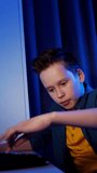 Portrait of teenager playing games. Happy boy sitting in his room and pushing the keyboard buttons while playing on PC. Blue evening light. Vertical video
