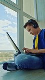 Happy boy in headphones with a laptop at home. School boy is sitting by the window and playing video games joyfully. Gaming concept. Vertical video