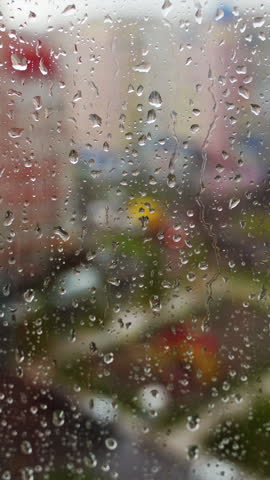 Wet window on blurred city background. Raindrops moving down on glass window at rainy weather. Water droplets on window pane. Vertical video